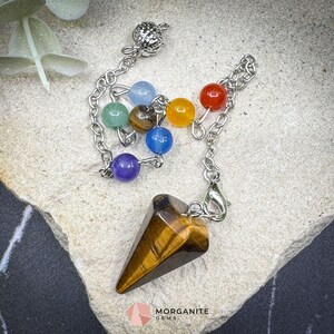 May include: A pendulum with a tiger's eye crystal point and a chain with colorful gemstone beads. The pendulum is a tool for divination and spiritual practices. The gemstones include blue, green, yellow, and red beads.
