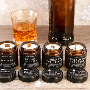 May include: Six amber glass candle jars with black lids and wooden wicks. The candles are labeled with the scents "Whiskey", "Dry Gin + Cypress", "Tobacco (The Noir 29)", and "Sandalwood + Bourbon".