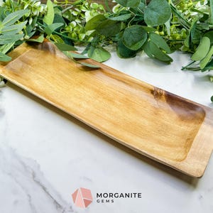 May include: A rectangular wooden serving tray with a light brown interior and a darker brown border. The tray is empty and sits on a white marble surface, with green foliage in the background. The tray is approximately 18 inches long.