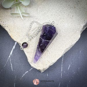 Może przedstawiać: Fioletowy wahadło z kryształu ametystu z srebrnym łańcuszkiem i małą fioletową koralikiem z ametystu. Wahadło leży na jasnoszarym kamiennym podłożu.