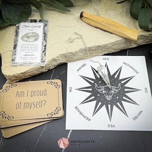 May include: A white crystal pendulum hangs over a black and white star-shaped chart with the words "Yes", "No", "Maybe", and "Rephrase" around the perimeter. The chart also features a bull's head in the center. A stack of brown cards with the question "Am I proud of myself?" is in the lower left corner. A small card with the words "Clear Quartz Power" is in the upper left corner.