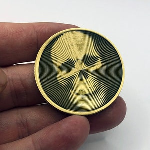 May include: A gold-colored metal coin with a black and white skull design. The skull is created using a spiral pattern.