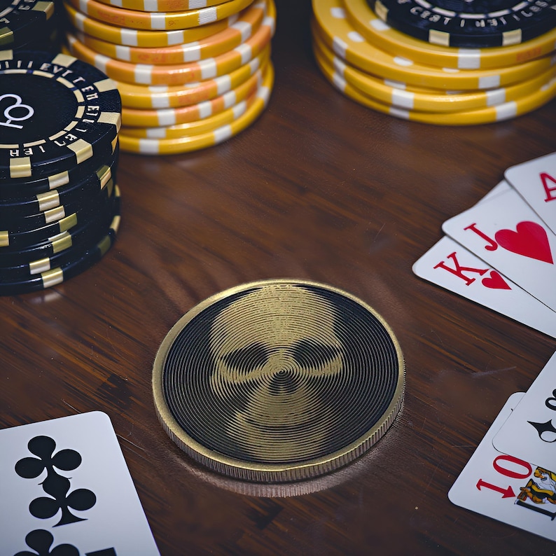 May include: A gold-colored poker chip with a skull design in the center. The chip is on a wooden table with playing cards and other poker chips.