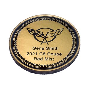 Op de afbeelding: Een ronde gouden munt met een zwarte rand en een zwart-wit geblokt vlaglogo. De tekst "Gene Smith 2021 C8 Coupe Red Mist" staat op de munt gedrukt.