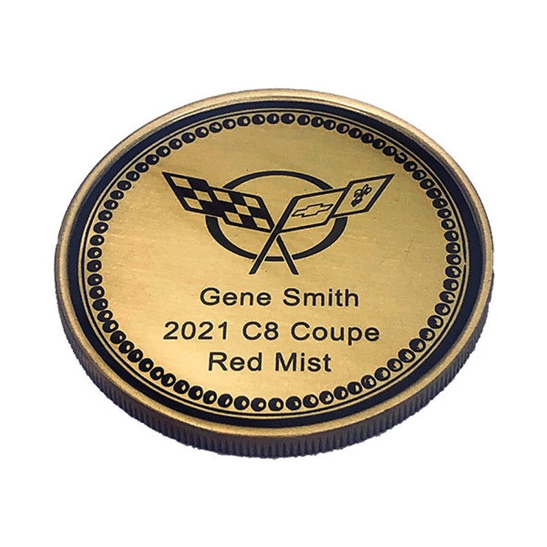 Op de afbeelding: Een ronde gouden munt met een zwarte rand en een zwart-wit geblokt vlaglogo. De tekst "Gene Smith 2021 C8 Coupe Red Mist" staat op de munt gedrukt.