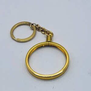 May include: A gold-colored key chain with a round coin holder. The coin holder is open and ready to hold a coin.