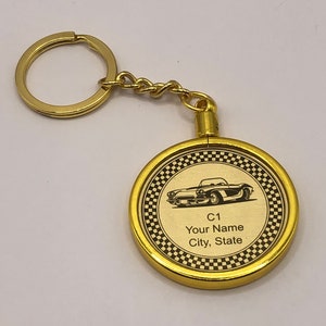 Op de afbeelding: Gouden sleutelhanger met een ronde metalen hanger met een zwart-wit geblokte rand. De hanger heeft een zwart-witte afbeelding van een klassieke auto met de tekst "C1 Your Name City, State".