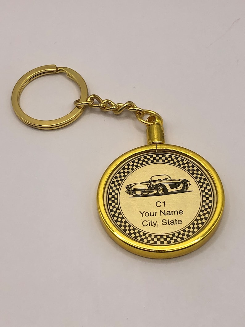Op de afbeelding: Gouden sleutelhanger met een ronde metalen hanger met een zwart-wit geblokte rand. De hanger heeft een zwart-witte afbeelding van een klassieke auto met de tekst "C1 Your Name City, State".