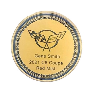 Op de afbeelding: Een ronde gouden munt met een blauwe rand. De munt heeft een logo met gekruiste vlaggen en de tekst "Gene Smith 2021 C8 Coup&eacute; Red Mist".