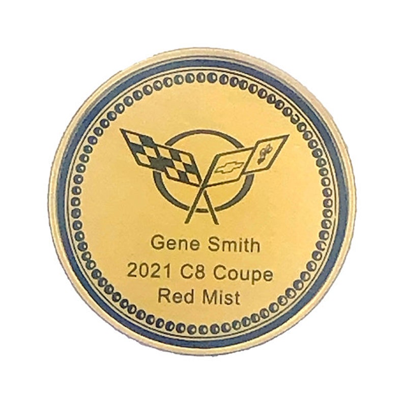 Op de afbeelding: Een ronde gouden munt met een blauwe rand. De munt heeft een logo met gekruiste vlaggen en de tekst "Gene Smith 2021 C8 Coup&eacute; Red Mist".
