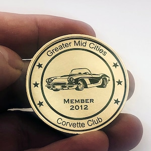 Op de afbeelding: Een gouden metalen munt met een zwarte omtrek en de tekst "Greater Mid Cities Corvette Club" rond de rand. Het midden van de munt heeft een zwart-witte illustratie van een klassieke Corvette met de tekst "MEMBER 2012" eronder.