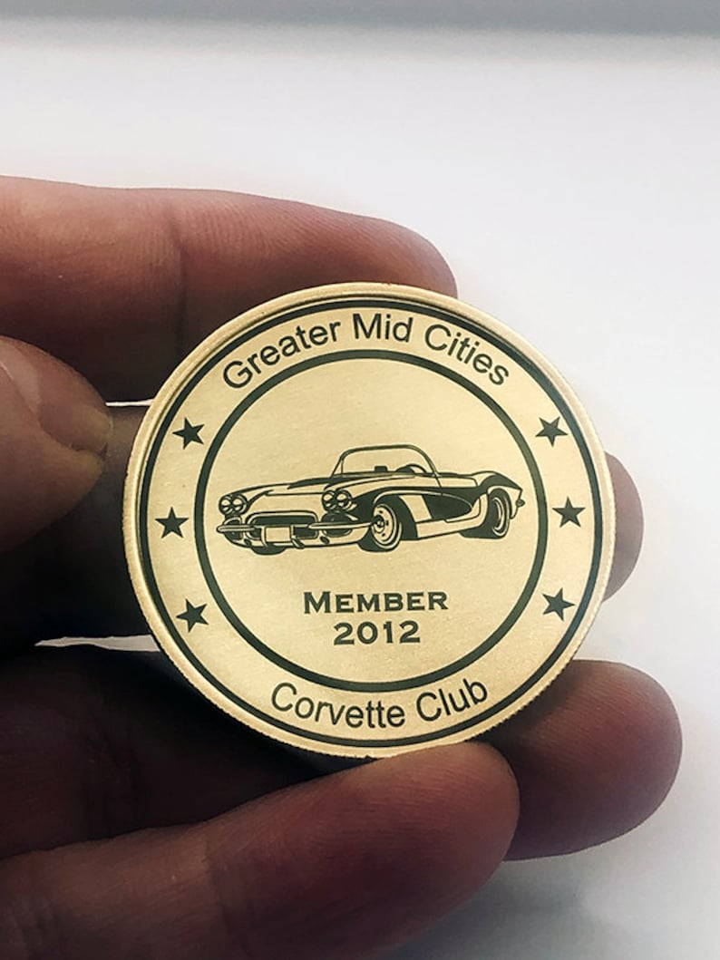 Op de afbeelding: Een gouden metalen munt met een zwarte omtrek en de tekst "Greater Mid Cities Corvette Club" rond de rand. Het midden van de munt heeft een zwart-witte illustratie van een klassieke Corvette met de tekst "MEMBER 2012" eronder.