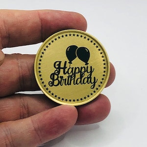 May include: Gold-colored oval coin with a black border of stars. The coin has a black inscription that reads "Happy Birthday" with two black balloons above the text.