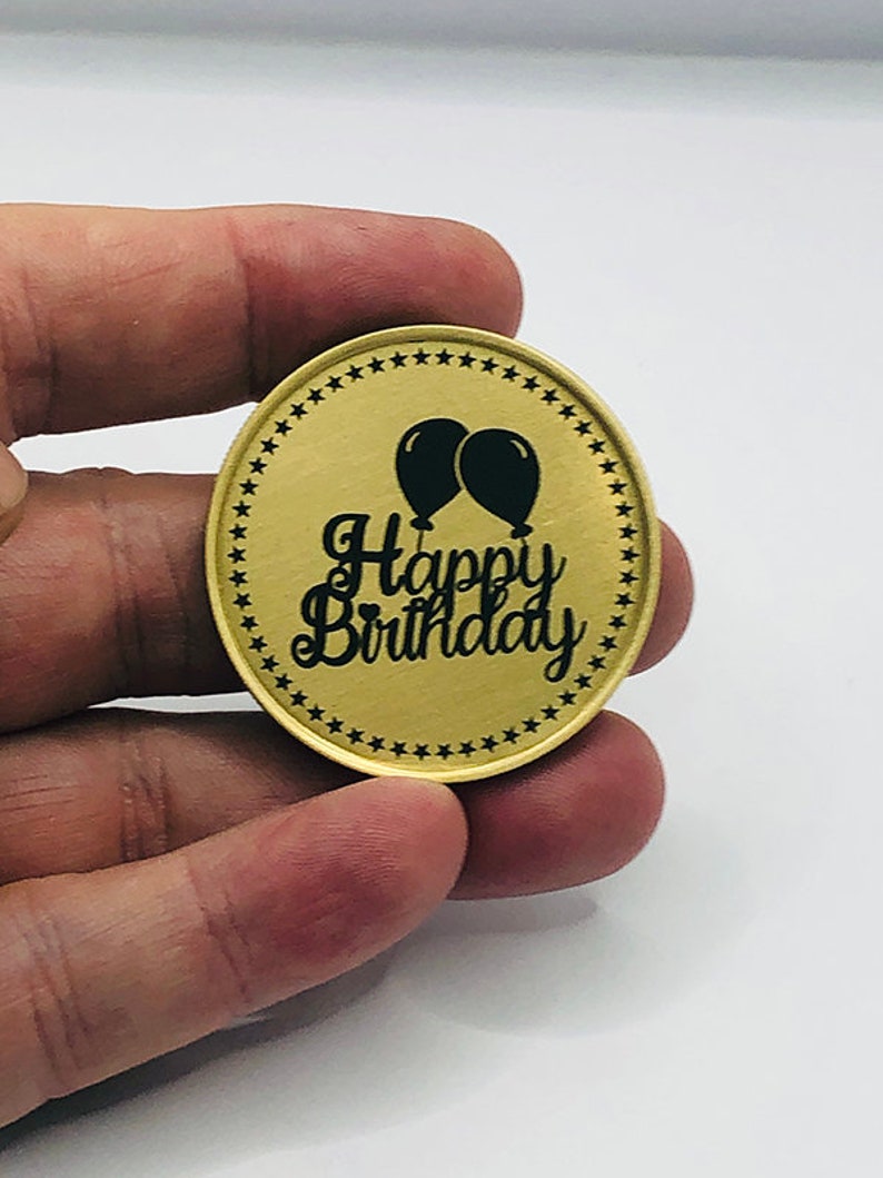May include: Gold-colored oval coin with a black border of stars. The coin has a black inscription that reads "Happy Birthday" with two black balloons above the text.