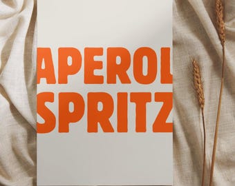 May include: A rectangular white print with the words "APEROL SPRITZ" in bold orange letters. The print is set against a neutral-toned fabric background, with two stalks of wheat on the right side.