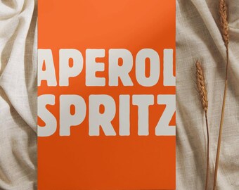 May include: An orange rectangular sign with the words "APEROL SPRITZ" in bold white letters. The sign is placed on a beige, textured fabric. Two stalks of wheat are positioned to the right of the sign.