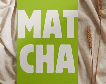 May include: A bright green rectangular sign with the word "MATCHA" in large, white, rounded letters. The sign is set against a neutral-toned, textured fabric background. Two stalks of wheat are placed to the right of the sign.