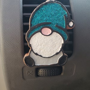 May include: A gnome-shaped car air freshener. The gnome has a teal glitter hat, a pink nose, and a white beard. The outline is black with a brown border. It hangs from a silver clip.