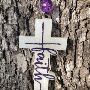May include: A white cross with the word "faith" written in purple glitter. The cross is hanging from a black cord with a purple glitter ball at the top.
