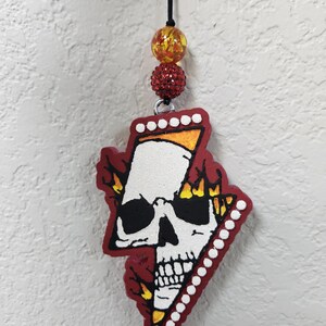 May include: A red and white skull-shaped ornament with a black string and decorative beads. The skull has a white face, black eyes, and a red border with flames. The ornament is approximately 3 inches in length.