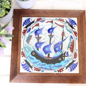 May include: A framed ceramic tile with a blue and white design of a ship with sails. The ship is surrounded by a border of red, blue, and white floral designs.