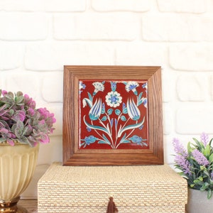 Puede incluir: Un azulejo de cerámica enmarcado con fondo rojo y diseño floral azul y blanco. El azulejo está rodeado por un marco de madera.