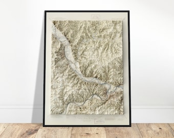 Puede incluir: Un mapa topográfico enmarcado con un marco negro. El mapa presenta un relieve detallado de un valle fluvial y el terreno circundante en tonos beige y marrón. El mapa se muestra en una pared blanca y un suelo de madera.