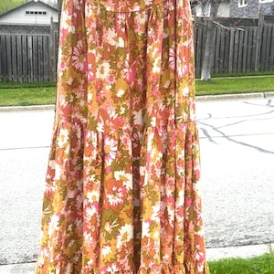May include: A long, floral-print dress in shades of orange, pink, and green hangs on a wooden hanger. The dress features a tiered skirt and spaghetti straps. The hanger has the words "WILLIAM PITT" and "HOTEL Chatham, Ont." printed on it.