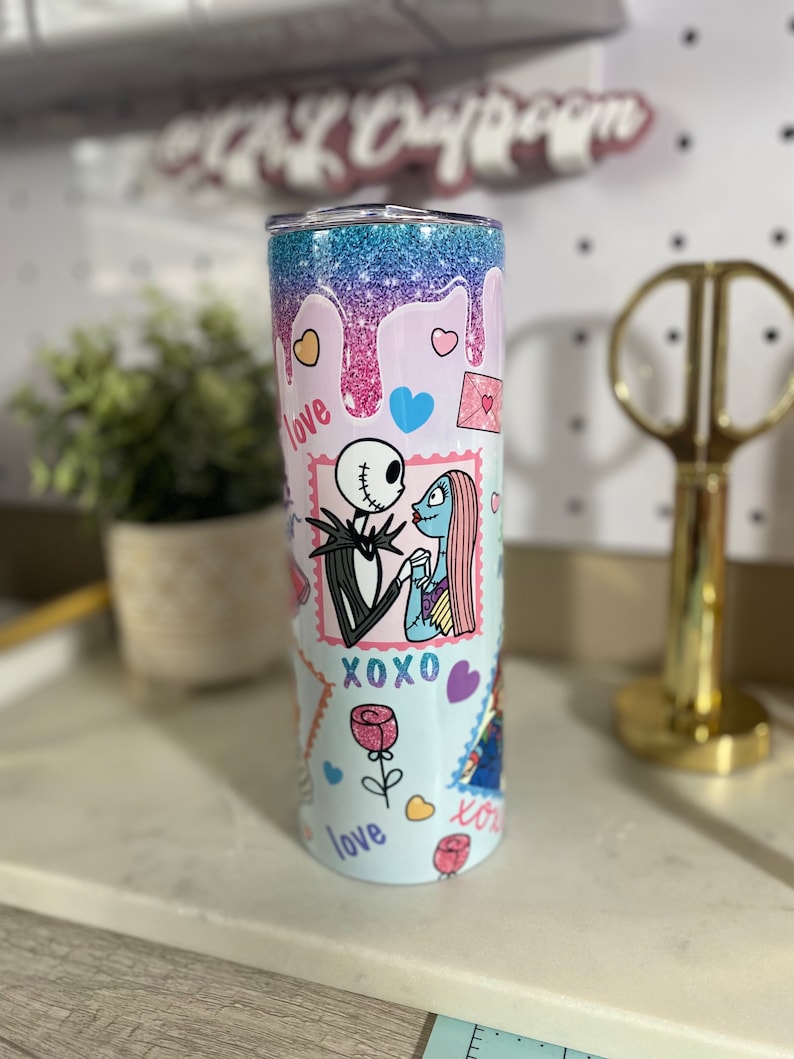 Puede incluir: Un vaso de acero inoxidable azul claro con un dise&ntilde;o de purpurina rosa, azul y morado. El vaso presenta un gr&aacute;fico de Jack Skellington y Sally de la pel&iacute;cula Pesadilla antes de Navidad. El gr&aacute;fico incluye las palabras "love", "xoxo" y "love" en una fuente rosa, azul y morada.