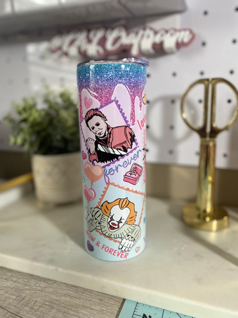 Puede incluir: Un vaso de glitter rosa y azul con fondo blanco. El vaso presenta un gr&aacute;fico de un personaje de la pel&iacute;cula "It" y un personaje de la pel&iacute;cula "Halloween". El texto "Now & Forever" est&aacute; impreso en el vaso.