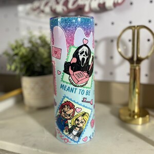 Puede incluir: Un vaso azul claro con acabado brillante. El vaso presenta un dise&ntilde;o gr&aacute;fico con personajes de pel&iacute;culas de terror, incluyendo Ghostface y Chucky. El texto "Meant to be" aparece en el vaso.