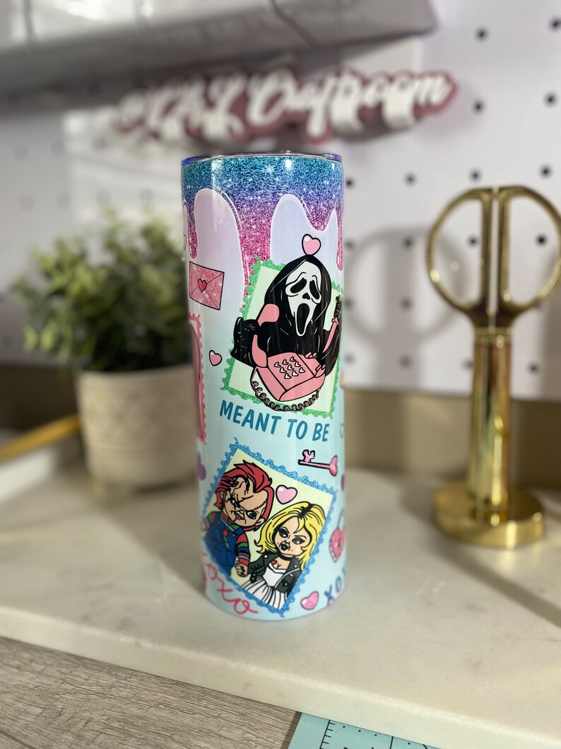Puede incluir: Un vaso azul claro con acabado brillante. El vaso presenta un dise&ntilde;o gr&aacute;fico con personajes de pel&iacute;culas de terror, incluyendo Ghostface y Chucky. El texto "Meant to be" aparece en el vaso.