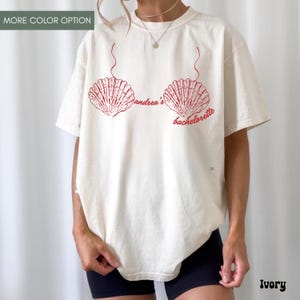 May include: Ivory-colored t-shirt featuring a red seashell bra design with the text "andrea's bachelorette". The shirt has a relaxed fit and short sleeves. The words "More Color Option" are at the top left.