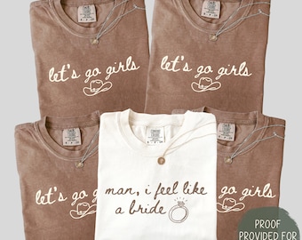 May include: Five t-shirts, four in brown and one in white, are displayed. The brown shirts have the text "let's go girls" with a cowboy hat graphic. The white shirt says "man, I feel like a bride" with a ring graphic. Each shirt has a necklace.