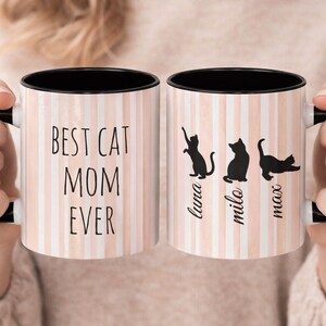 May include: Two white ceramic mugs with black handles and rims. The mugs have a pink and white striped pattern. One mug says "BEST CAT MOM EVER" in black text. The other mug has black cat silhouettes and the names "luna", "milo", and "max".