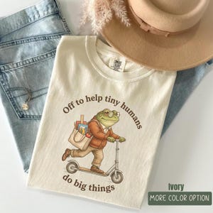 May include: An ivory t-shirt featuring a graphic of a green frog wearing glasses, a brown cardigan, and tan trousers while riding a scooter and carrying a tote bag. The text reads, Off to help tiny humans do big things. The shirt is paired with blue jeans.
