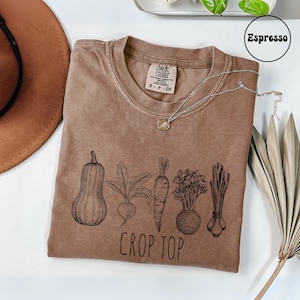May include: Espresso-colored crop top with a crew neck, featuring a black illustration of vegetables: a squash, beet, carrot, celery root, and scallions. The words "CROP TOP" are printed below the vegetables. A silver necklace rests on the shirt.