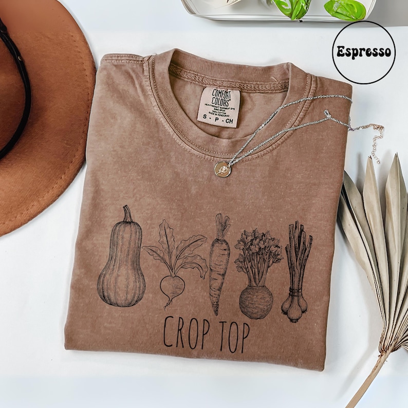 May include: Espresso-colored crop top with a crew neck, featuring a black illustration of vegetables: a squash, beet, carrot, celery root, and scallions. The words "CROP TOP" are printed below the vegetables. A silver necklace rests on the shirt.
