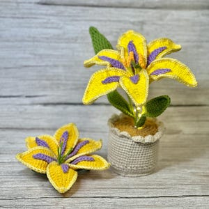 May include: Two crocheted yellow lilies with purple centers in a crocheted pot. The lilies are in a light wood background.