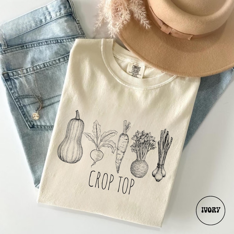 May include: Ivory-colored crop top with a black and white illustration of various vegetables, including a squash, beet, carrot, and leeks. The words "CROP TOP" are printed below the vegetables. The shirt is paired with blue jeans and a tan hat.