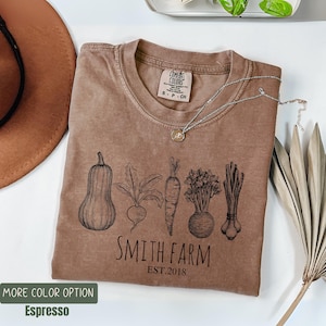 May include: Espresso-colored t-shirt with a graphic of vegetables, including a squash, beet, carrot, and leeks. The text "SMITH FARM EST.2018" is printed below the vegetables. A silver necklace with a pendant rests on the shirt.