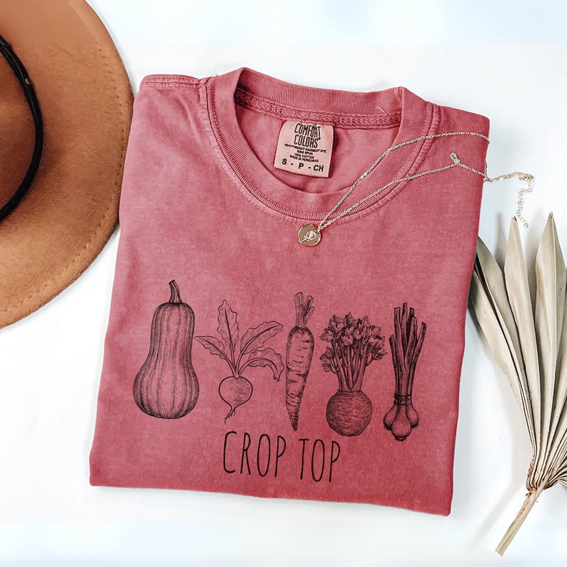 May include: A dusty rose crop top with a black graphic of vegetables including a butternut squash, beet, carrot, and leeks. The words "CROP TOP" are printed below the vegetables. A silver necklace with a small pendant is on the shirt.
