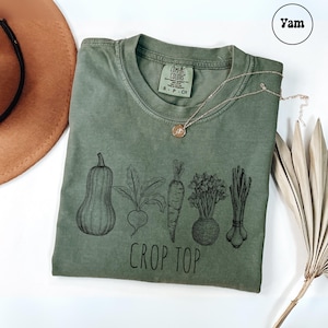 May include: Olive green t-shirt with a black graphic of various vegetables and the words "CROP TOP". A gold necklace with a pendant rests on the shirt. A brown hat and dried plant are in the background.