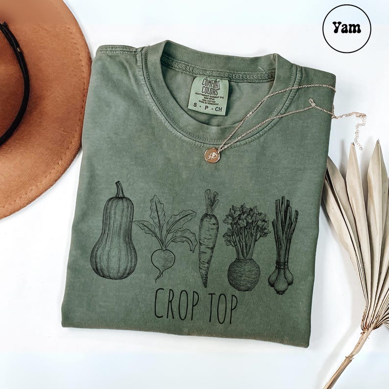 May include: Olive green t-shirt with a black graphic of various vegetables and the words "CROP TOP". A gold necklace with a pendant rests on the shirt. A brown hat and dried plant are in the background.