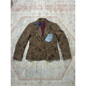 May include: A brown blazer featuring an all-over dark floral pattern, a deep burgundy inner lining, and a light blue denim patch pocket on the left chest. The garment has a notched lapel, long sleeves, and two dark buttons for front closure.