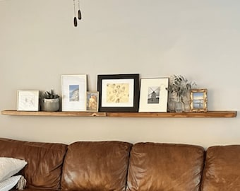 May include: A wooden floating shelf with a brown finish, mounted on a white wall. The shelf is decorated with framed pictures, a small potted plant, and decorative items. A brown leather sofa is in the foreground.