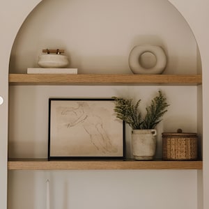 May include: A white ceramic vase with a circular opening sits on a wooden shelf with a framed drawing of a nude figure, a small wooden box, and a potted plant. The shelf is part of a built-in arched alcove.