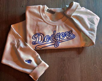 May include: Cream-colored sweatshirt with the word "Dodgers" in blue script across the chest. The left sleeve cuff features a blue embroidered logo. The sweatshirt is folded on a wooden surface.