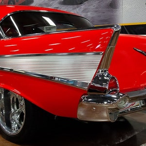 May include: A red 1957 Chevrolet Bel Air with chrome accents and a white interior. The car is in a showroom setting with a watercolor effect.