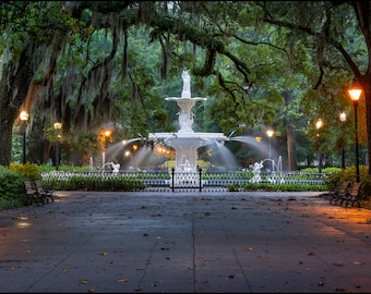 May include: A white fountain with water spraying from the top, surrounded by lush green trees and a black wrought iron fence. The fountain is located in a park with a paved walkway leading to it. There are streetlights lining the walkway, casting a warm glow on the scene.